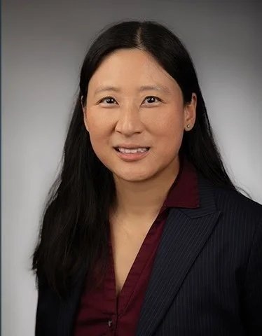 A woman with long black hair wearing a dark navy blazer over a maroon blouse, smiling slightly, against a neutral gray background.