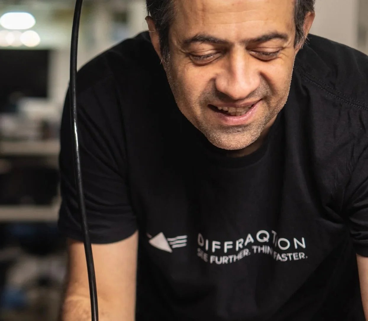 A man with gray hair and stubble smiling, wearing a black T-shirt with the word 'Differation' and the slogan 'Go Further. Think Faster.' in a blurred indoor setting.